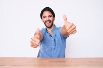 Handsome hispanic man wearing casual clothes sitting on the table approving doing positive gesture with hand, thumbs up smiling and happy for success. winner gesture.