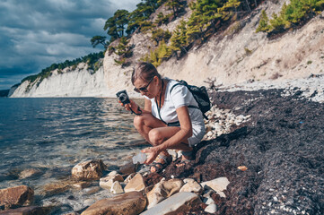 Elderly senior traveling backpacker mature woman tourist walking taking photos on coast background of sea, stones, rocks, blue sky. Retired people summer holiday vacation, active lifestyle concept