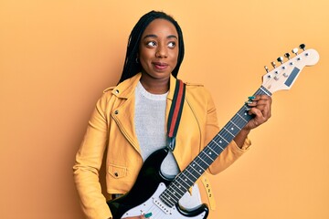 Young african american woman playing electric guitar smiling looking to the side and staring away thinking.