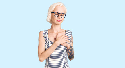 Young blonde woman with tattoo wearing casual clothes and glasses smiling with hands on chest with closed eyes and grateful gesture on face. health concept.