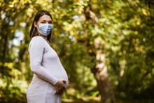 Portrait of pregnant woman in park in autumn. She is wearing protective face mask. Covid-19 pandemic concept.