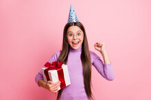 Photo Of Delighted Little Kid Girl Hold Gift Box Raise Fists Wear Purple Brithday Cone Isolated On Pastel Color Background