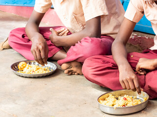 Unidentified poor classmates children with uniforms sitting on the floor outdoors, eating with their right hand some rice with masala. Lunch time