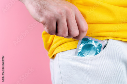 A man in white jeans pulls a condom out of his pocket. Hand close-up. Pink background. Concept of protection against sexually transmitted infections. Copy space