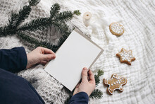 Christmas Festive Still Life. Little Child Hands Holding Blank Greeting Card. Invitation Mockup On Knitted Sweater. Gingerbread Cookies And Fir Branches On White Muslin Blanket. Selective Focus.
