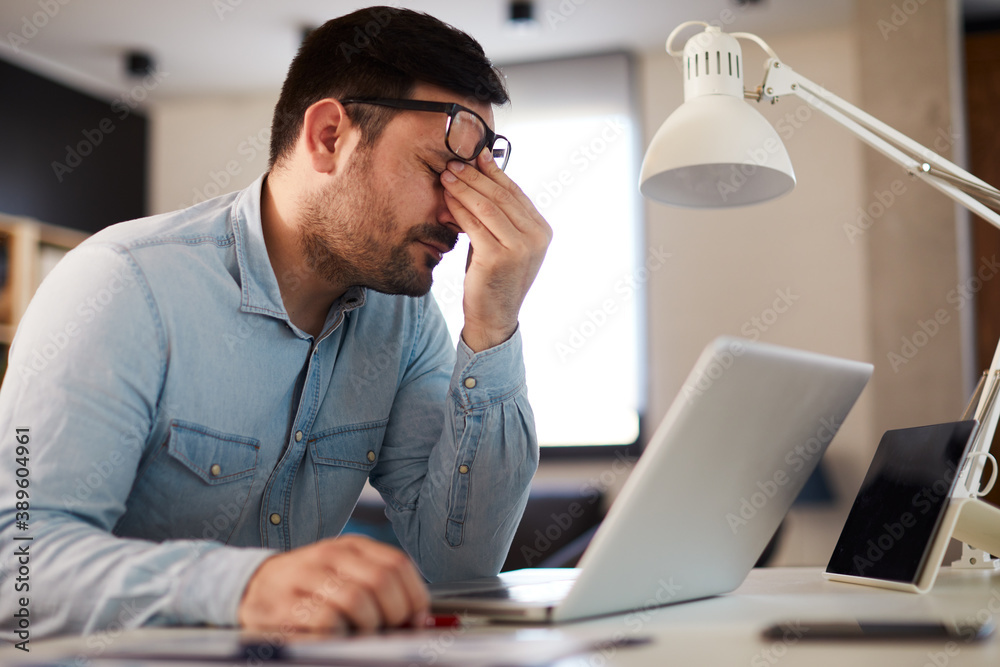  Young man has a headache at work while working on a laptop - obrazy, fototapety, plakaty 