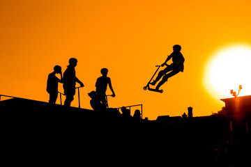 Wall Mural - Unrecognizable teenage boy silhouette showing high jump tricks on scooter against orange sunset sky at skatepark. Sport, freestyle, extreme, youth, urban culture, outdoor activity concept