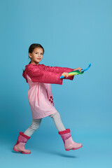 Wall Mural - happy funny child girl with multicolored umbrella in pink rain coat and rubber boots on blue background.