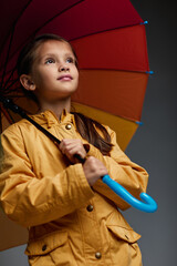 Wall Mural - Cheerful child girl with yellow and red umbrella in yellow rain coat on gray background.