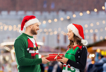 Wall Mural - people and holidays concept - happy couple in santa hats and ugly sweaters with gift box over christmas market background