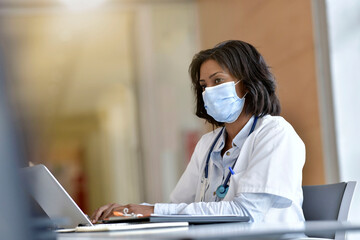 Wall Mural - Woman doctor working in office with laptop computer; wearing face mask