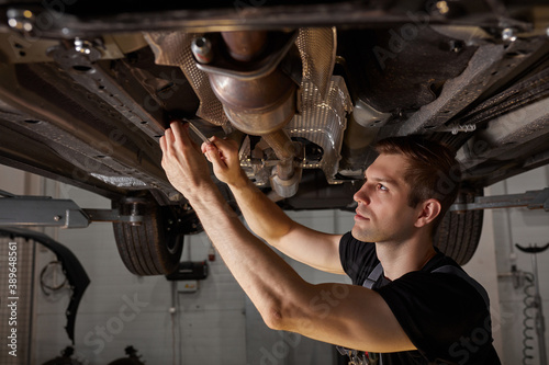young male car mechanic in uniform checking car in automobile service with lifted vehicle, handsome hardworking guy working under car condition on lifter. automotive car repair concept