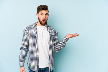 Wall Mural - Young caucasian man isolated on blue background impressed holding copy space on palm.
