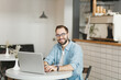 © ViDi Studio - Smiling man in eyeglasses sitting alone at table in coffee shop cafe working or studying on laptop pc computer relaxing in restaurant during free time indoor. Freelance mobile office business concept.