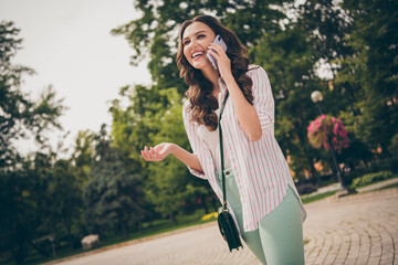 Wall Mural - Photo portrait of pretty girl talking on cellphone laughing walking in green city park wearing casual outfit