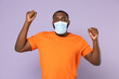 © ViDi Studio - Joyful young african american man 20s in casual orange t-shirt sterile face mask to safe from coronavirus virus covid-19 doing winner gesture isolated on pastel violet background, studio portrait.