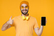 © ViDi Studio - Smiling young man in casual t-shirt headphones eyeglasses hat standing showing thumb up hold mobile phone with blank empty screen mock up copy space isolated on yellow background, studio portrait.