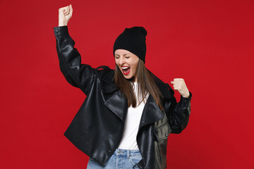 Wall Mural - Overjoyed happy young brunette woman wearing casual black leather jacket white t-shirt hat clenching fists doing winner gesture keeping eyes closed isolated on bright red background, studio portrait.