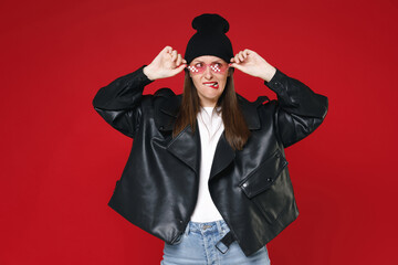 Wall Mural - Displeased puzzled perplexed young brunette woman 20s in casual black leather jacket white t-shirt hat eyeglasses biting lips looking aside isolated on bright red colour background studio portrait.