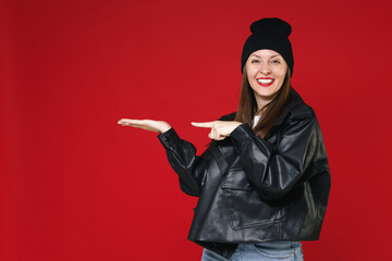 Wall Mural - Pretty smiling young brunette woman 20s wearing black leather jacket white t-shirt hat pointing index finger hand aside on mock up copy space isolated on bright red colour background studio portrait.