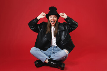 Wall Mural - Full length of excited surprised young brunette woman 20s in casual black leather jacket white t-shirt hat sitting keeping mouth open looking camera isolated on bright red background studio portrait.