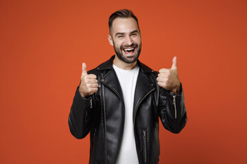 Excited laughing cheerful young bearded man 20s wearing basic white t-shirt, black leather jacket standing showing thumb up looking camera isolated on bright orange colour background, studio portrait.