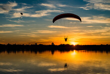 Two Powered Paragliders Flying Over The Lake During Sunset. Silhouetted Paramotor Pilot With Reflection In Water