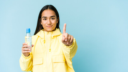 Young asian woman holding a bottle of water isolated on blue background showing number one with finger.