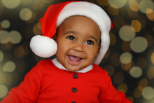 Cute Little African American Baby Wearing Santa Hat Against Blurred Lights On Dark Background. Christmas Celebration