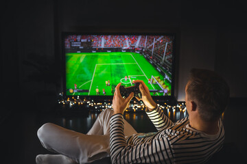 Young hipster man playing video game football soccer on console. gamer guy with gamepad controller holding Wireless joystick sitting on pouf at night. Back view. Fun entertainment at home isolation.