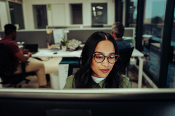 Wall Mural - Multi-ethnic female entrepreneur wearing spectacles working on business report typing on desktop computer sitting in office 