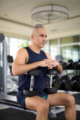 Mature handsome Persian man exercising with dumbbells and sitting at the gym