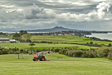 AUCKLAND, NEW ZEALAND - Mar 28, 2019: Lawn Mower Red Tractor Working In Macleans Park With Hauraki Gulf Islands In Background
