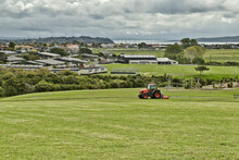 AUCKLAND, NEW ZEALAND - Mar 28, 2019: Lawn Mower Red Tractor Working In Macleans Park With Hauraki Gulf Islands In Background
