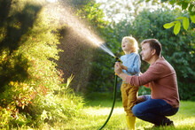 Funny Little Boy With His Father Watering Plants And Playing With Garden Hose In Sunny Backyard. Preschooler Child Having Fun With Spray Of Water.