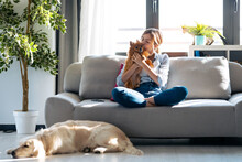 Attractive Young Woman Kissing Her Little Cute Dog While Sitting In Couch With Her Dogs And Cat In Living Room At Home.