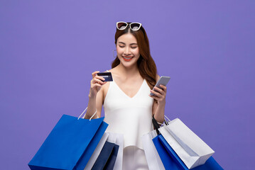 Portrait of smiling Asian woman purchasing online through phone using credit card while carrying shopping bags isolated on purple background