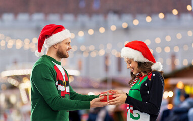 Wall Mural - people and holidays concept - happy couple in santa hats and ugly sweaters with gift box over christmas market background