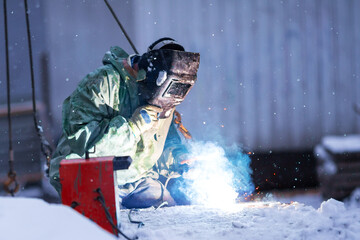 Welder in protective clothing and special protective mask performs welding work in winter.