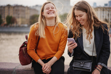 two beautiful young woman friends walking together around the city, using a smartphone and having fu