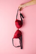 © fotoduets - Young adult woman hand holding black red bra on light pink background. Pastel color. Daily underwear. Closeup. Front view.