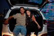 © Svitlana - A group of two young casually dressed friends of different genders and nationalities smiling sitting in an opened car trunk outside on a parking site with a led screen behind
