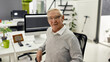 © Svitlana - Portrait of aged man, senior intern in wireless earphones smiling at camera while sitting at desk, working on pc in modern office