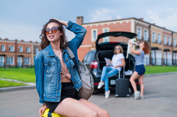 Three girlfriends go on a road trip. Young woman posing with a suitcase. Two girls are sitting on the trunk of a car and watching a map.