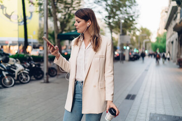 Youthful generation Z with takeaway coffee to go using smartphone application for searching media songs and listen on leisure, hipster girl in electronic earphones standing at urban street and phoning