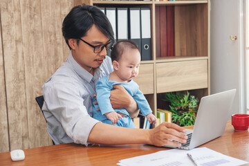 Father working online with a laptop holding baby son at home office. Single dad with him child. Asian man working from home, while in quarantine isolation during Covid-19 crisis.