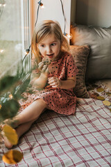 beautiful little girl sits on the windowsill and eats cookies. new year's interior and garlands. chr