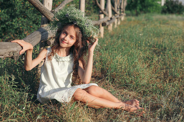 Cute little girl wearing wreath made of beautiful flowers near wooden fence outdoors on sunny day