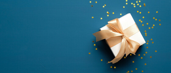 white gift box with golden ribbon bow on blue background with confetti. christmas present, valentine