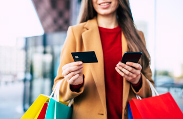 Wall Mural - Beautiful trendy young woman with many colorful shopping bags in good mood with smart phone and credit card while walking in the mall during black friday